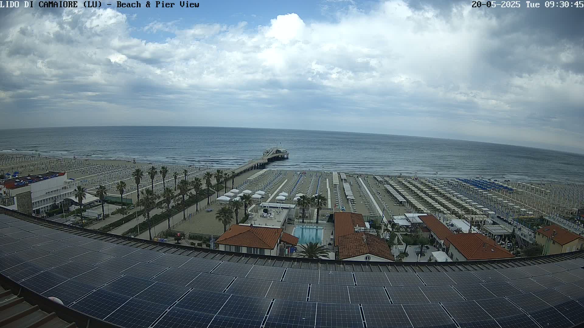 A beach with numerous umbrellas and a pier extends into a calm sea under a partly cloudy sky.