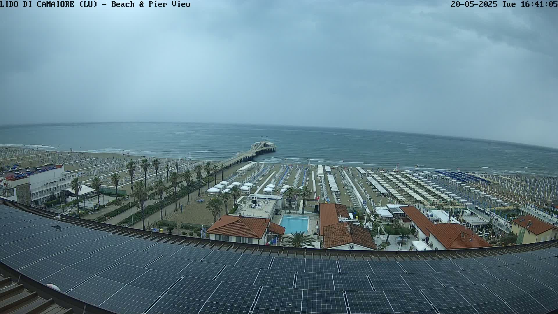 A beach with numerous umbrellas and a pier extends into a calm, overcast ocean, viewed from above a solar panel array on a building.