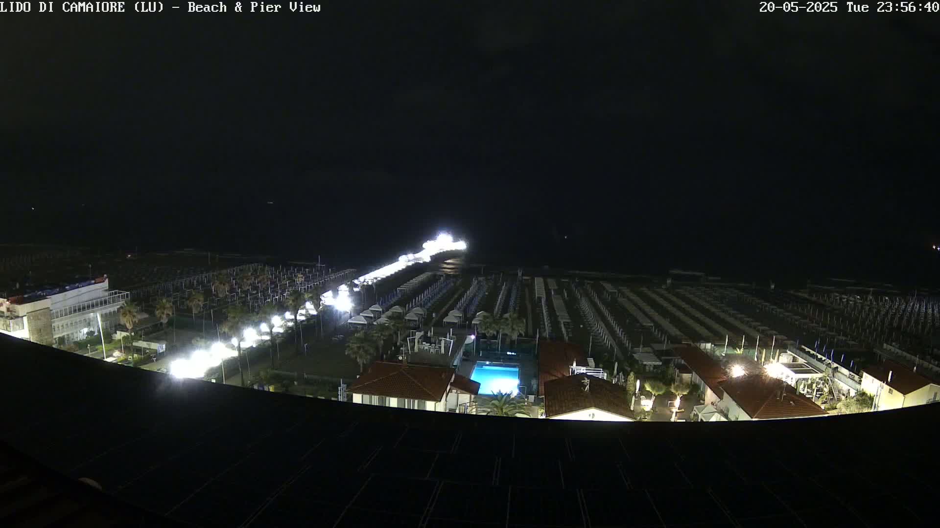 A nighttime, high-angle view of a beach at night, showing rows of beach umbrellas and chairs, a pier with lights, a building with a pool, and dark water in the distance.