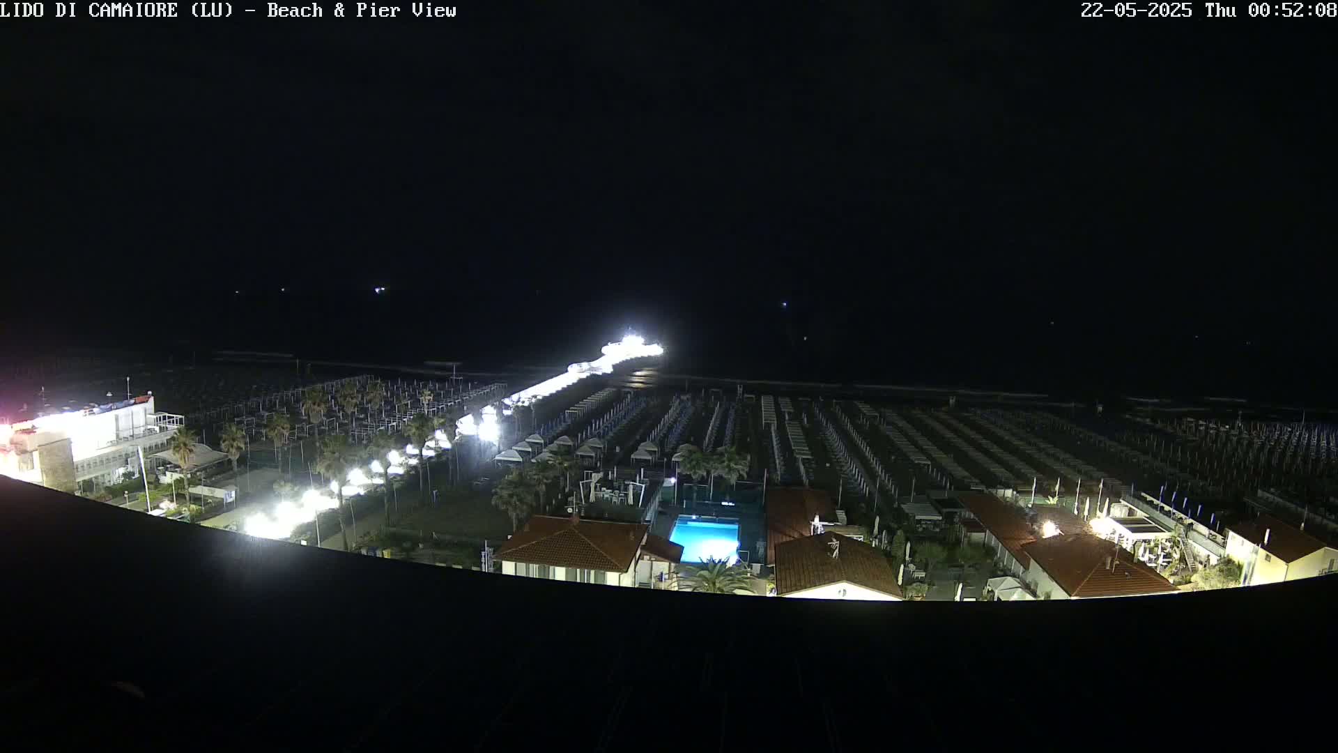 A nighttime, high-angle view shows a brightly lit pier extending from a beach lined with rows of beach umbrellas and buildings, all under a dark sky.
