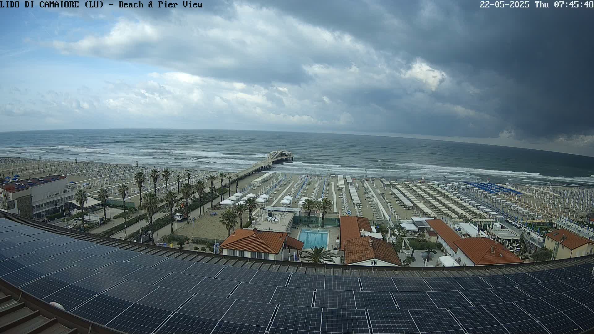 A beach with rows of umbrellas and lounge chairs under a cloudy sky, viewed from above a rooftop equipped with solar panels, showing a pier extending into a somewhat choppy sea.