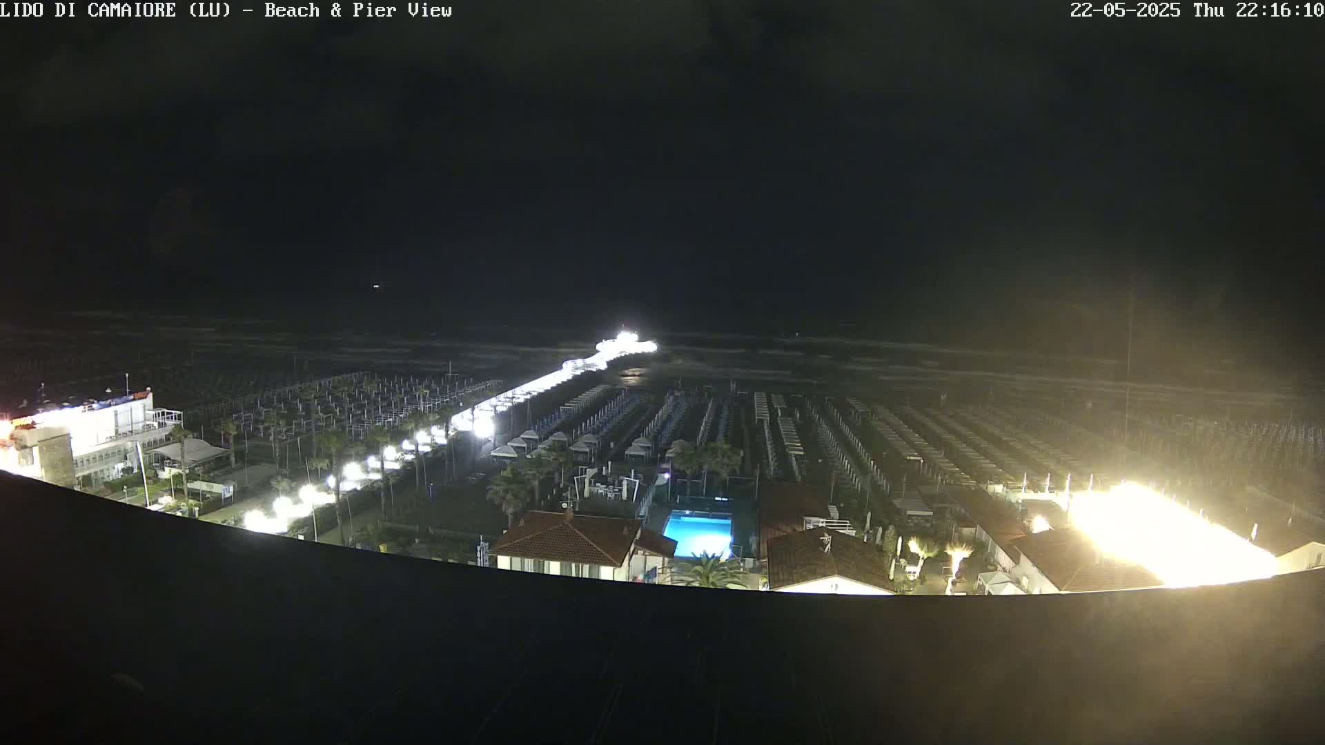 A nighttime, aerial view of a beach with rows of beach umbrellas and a pier extending into a dark ocean under a mostly clear sky.