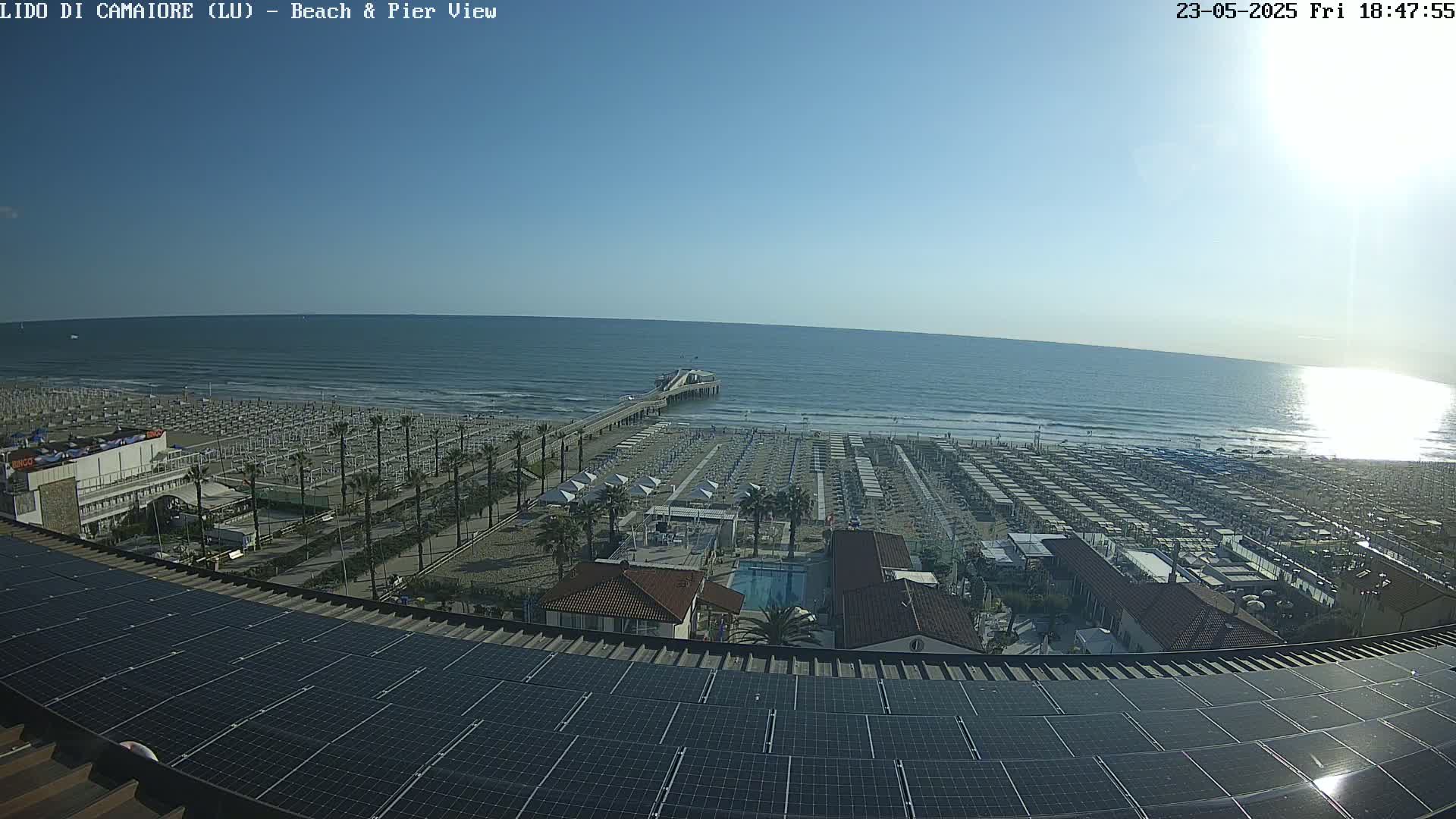 A sunny beach scene with calm ocean waves, numerous beach umbrellas, a pier extending into the water, and a building with solar panels on its roof.
