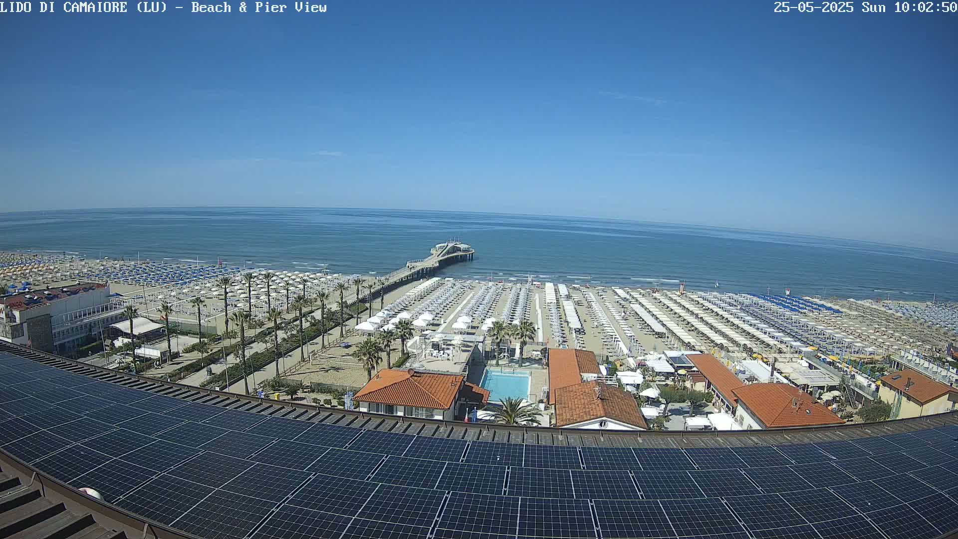 A beach with many umbrellas and a pier extends into a calm, blue ocean under a clear, sunny sky, viewed from above a rooftop covered in solar panels.