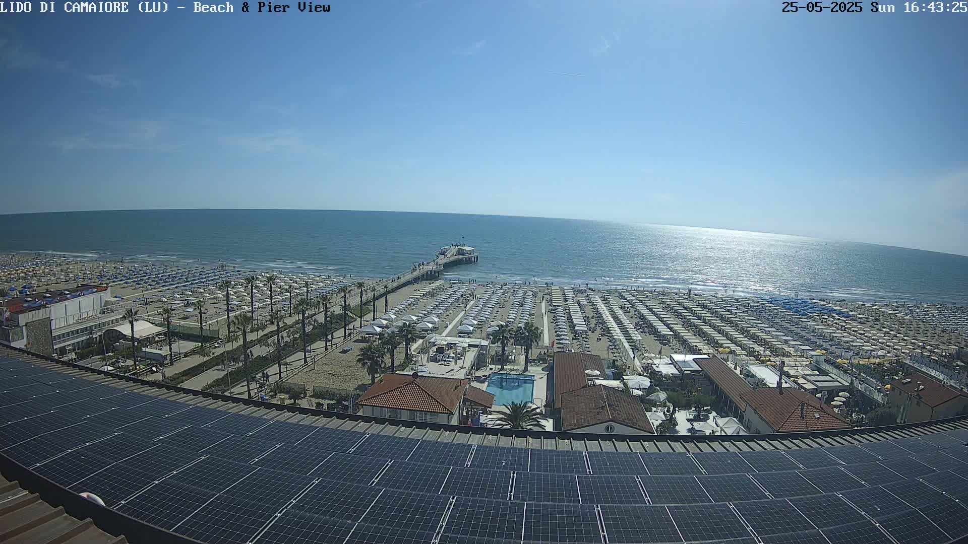 A sunny day overlooking a beach with numerous umbrellas, a pier extending into a calm sea, and a solar panel array on a rooftop.