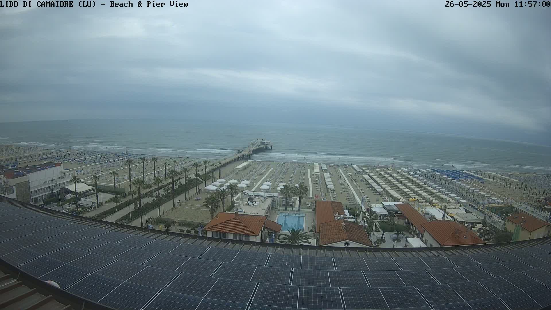 A beach with rows of umbrellas and a pier extends into a calm, overcast ocean, viewed from a rooftop with solar panels.