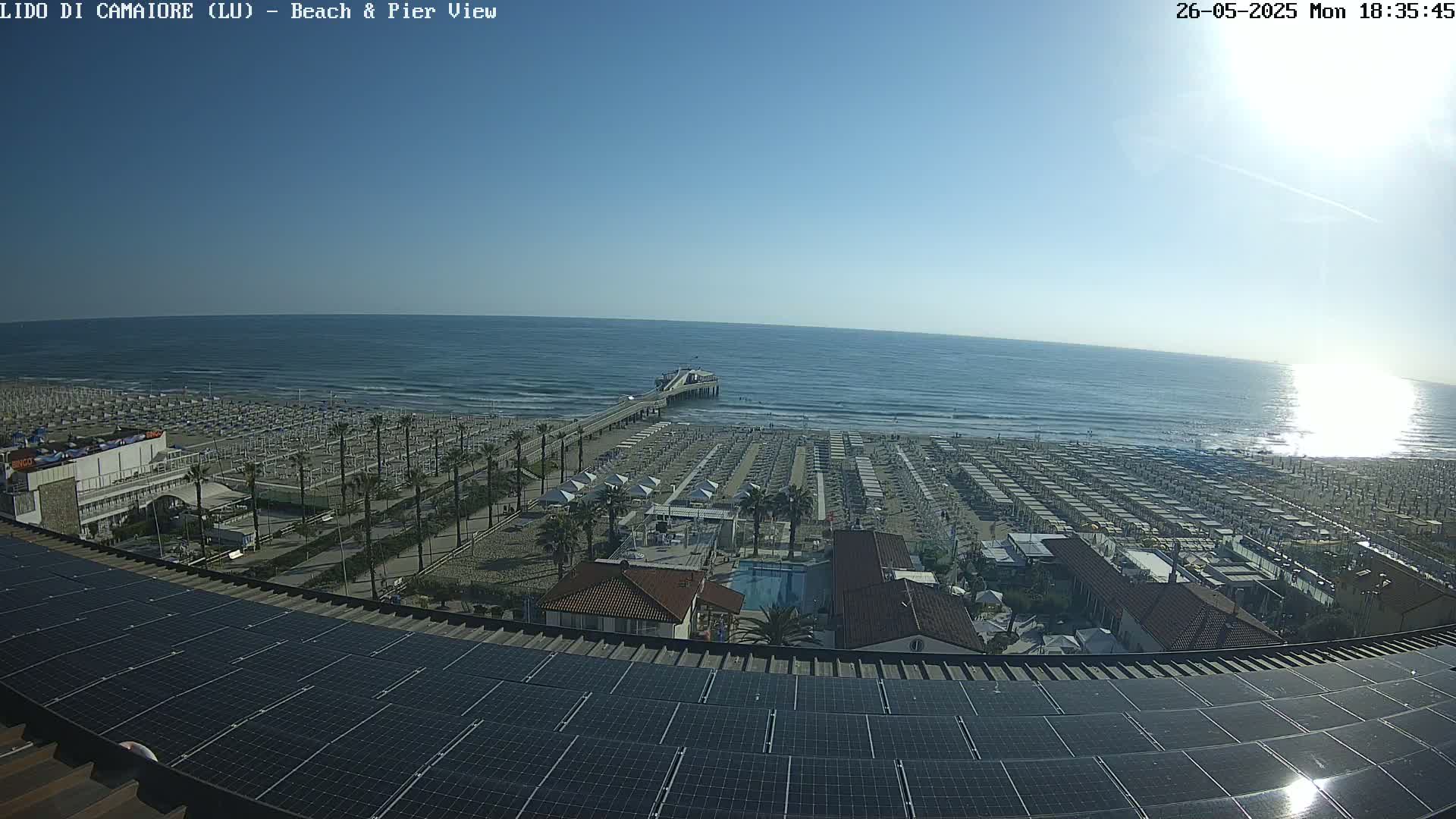 A beach with numerous umbrellas and a pier extends into a calm ocean under a clear, sunny sky, viewed from above a solar panel-covered roof.