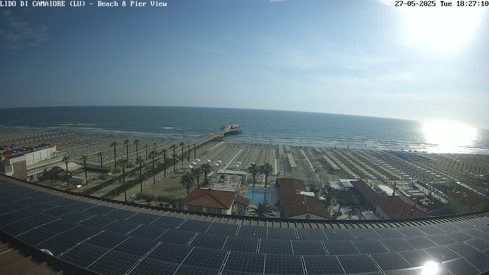 A beach with rows of umbrellas and a pier extends into a calm, sunny ocean, viewed from above a rooftop covered in solar panels.