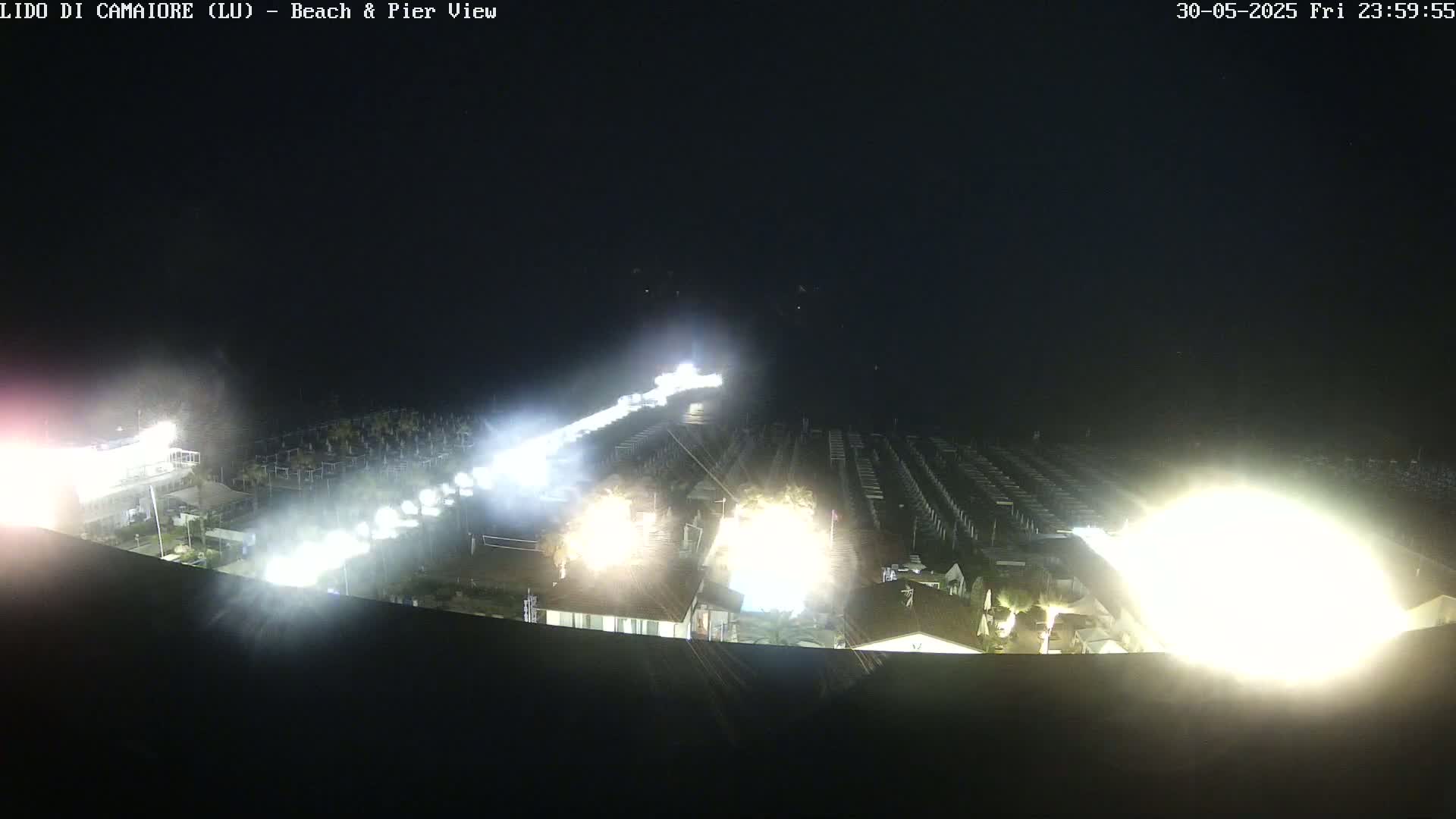 Bright lights illuminate a beach at night, with rows of beach structures visible under the strong light.