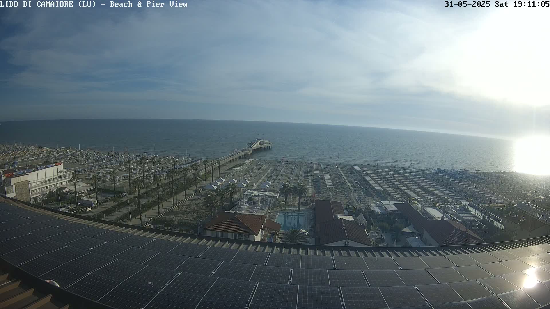 A mostly sunny beach scene is visible from above, showing a long pier extending into a calm ocean, numerous beach umbrellas lining the sand, and buildings and solar panels in the foreground.