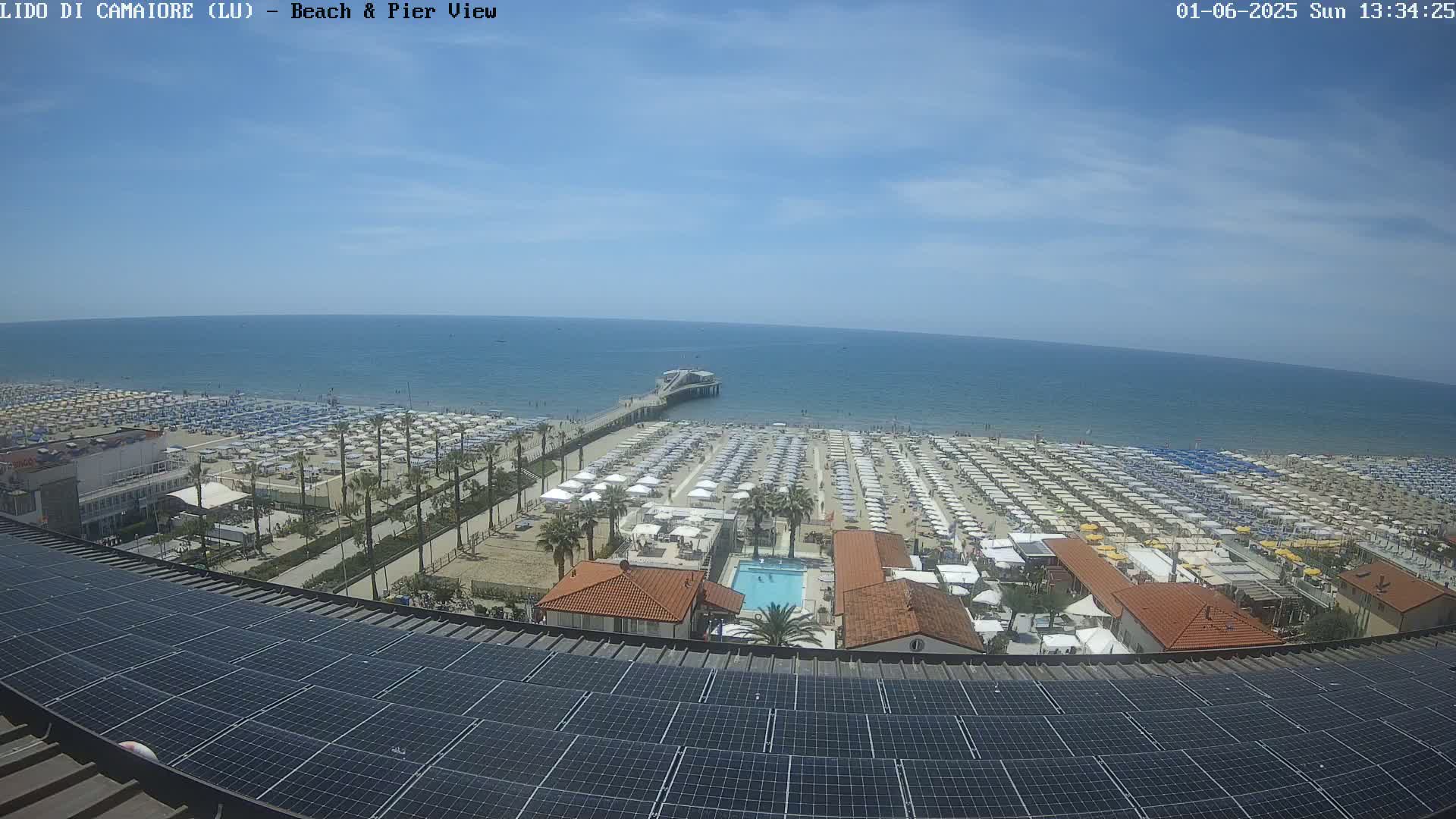 A sunny day shows a beach crowded with umbrellas and a pier extending into a calm ocean, viewed from above a rooftop covered in solar panels.