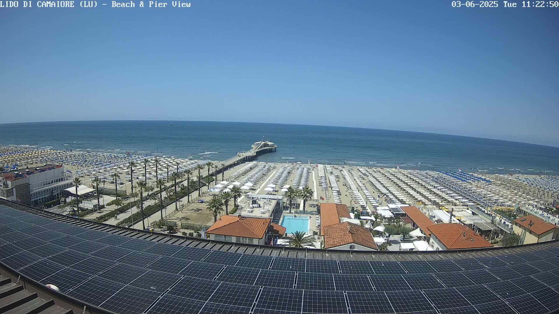A beach with numerous umbrellas and a pier extending into a calm, blue ocean is viewed from above a rooftop covered in solar panels on a sunny day.