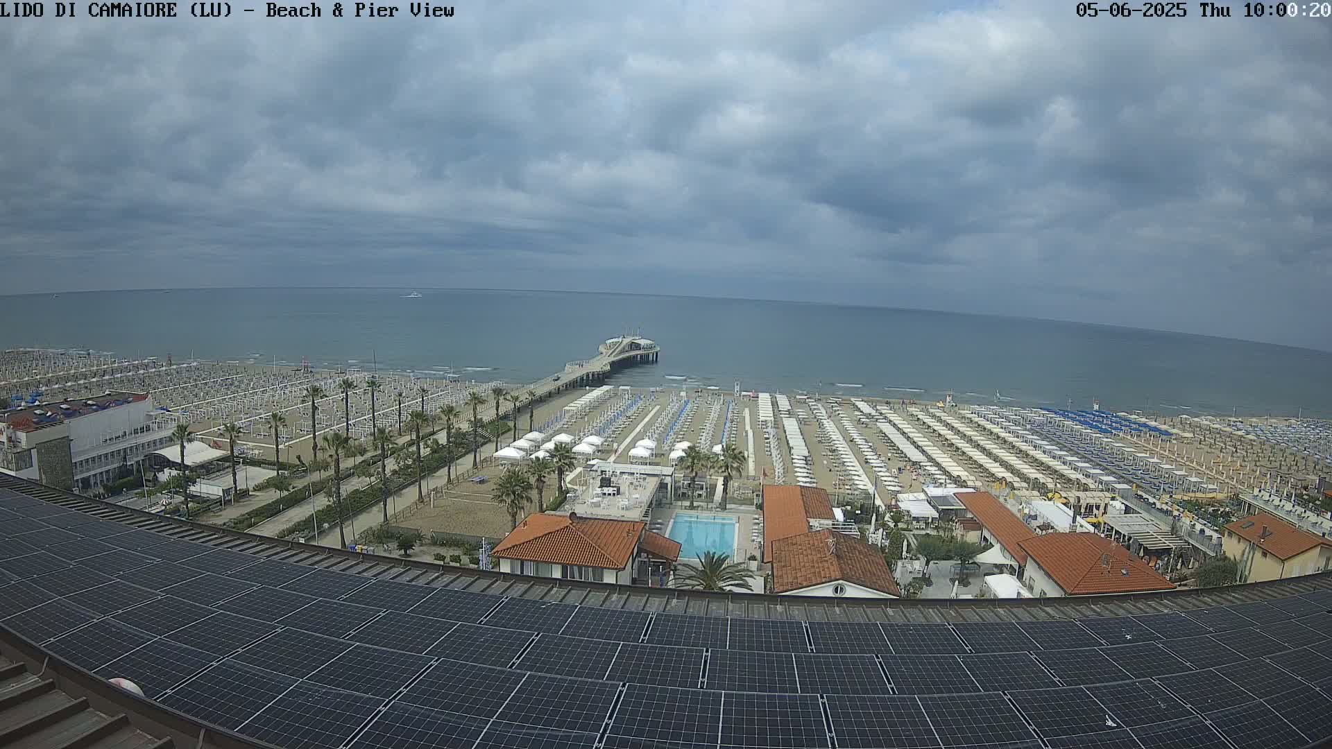 A beach with numerous umbrellas and a pier extending into a calm sea under a cloudy sky is viewed from above a solar panel array.