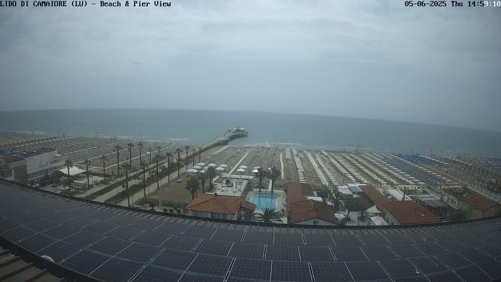 An expansive beach with numerous umbrellas and a pier extending into a calm, overcast ocean is viewed from a rooftop covered in solar panels.