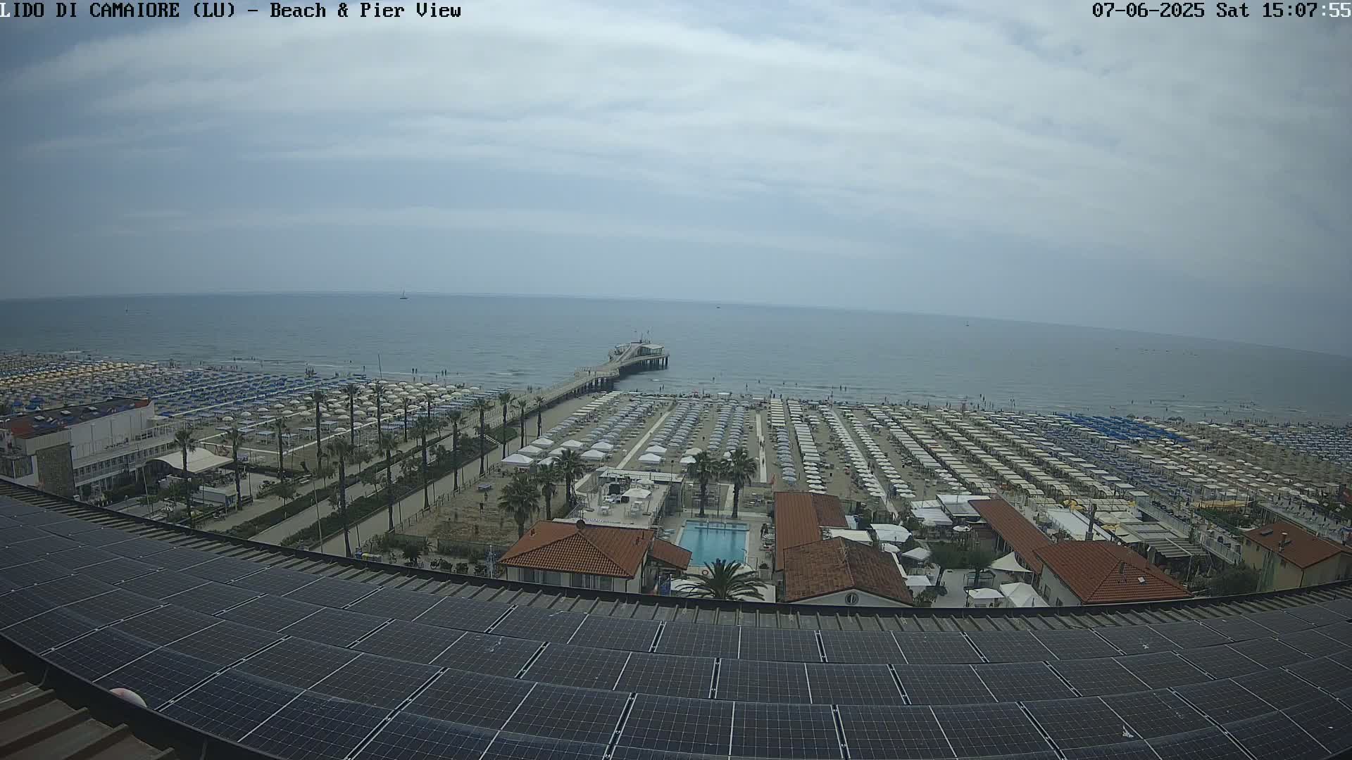 A beach with numerous umbrellas and a pier extends into a calm ocean under a partly cloudy sky, viewed from above a rooftop covered in solar panels.