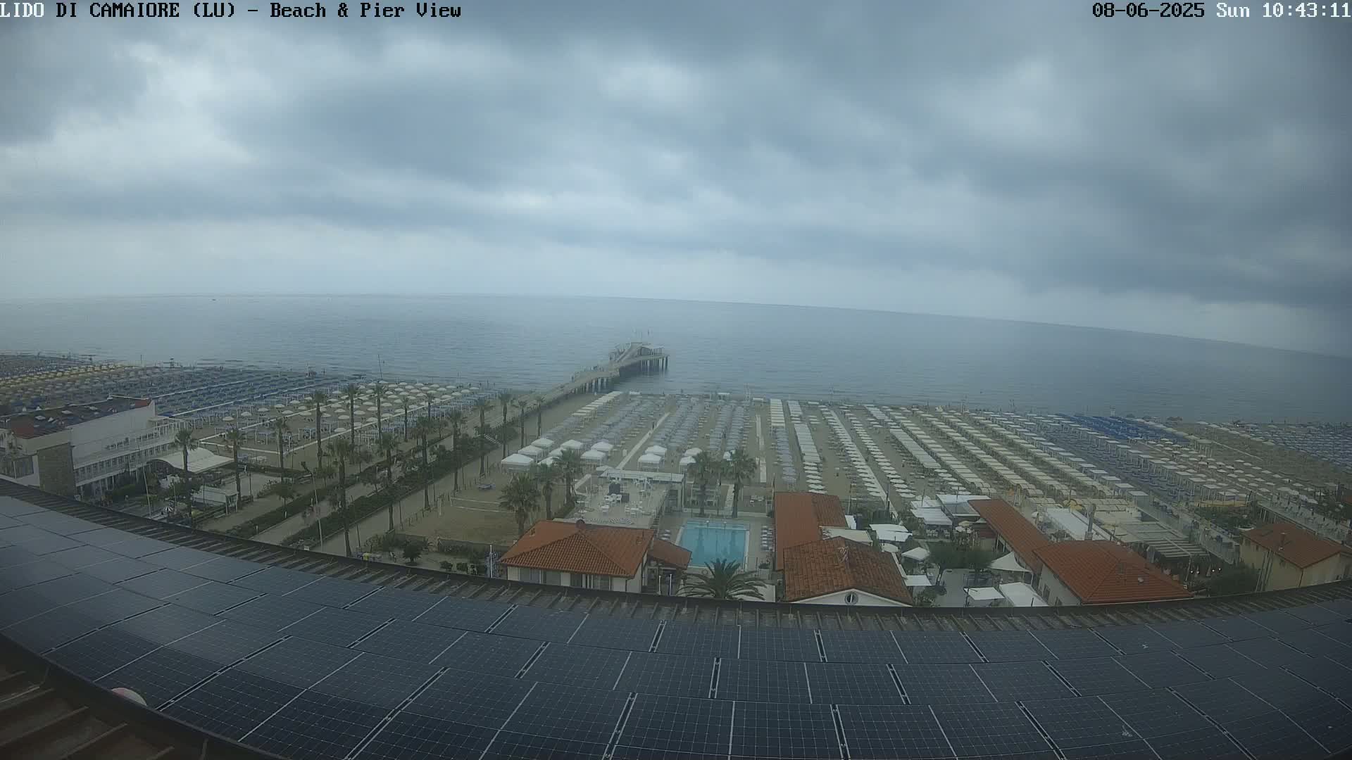 A beach with rows of umbrellas and a pier extends into a calm sea under an overcast sky.