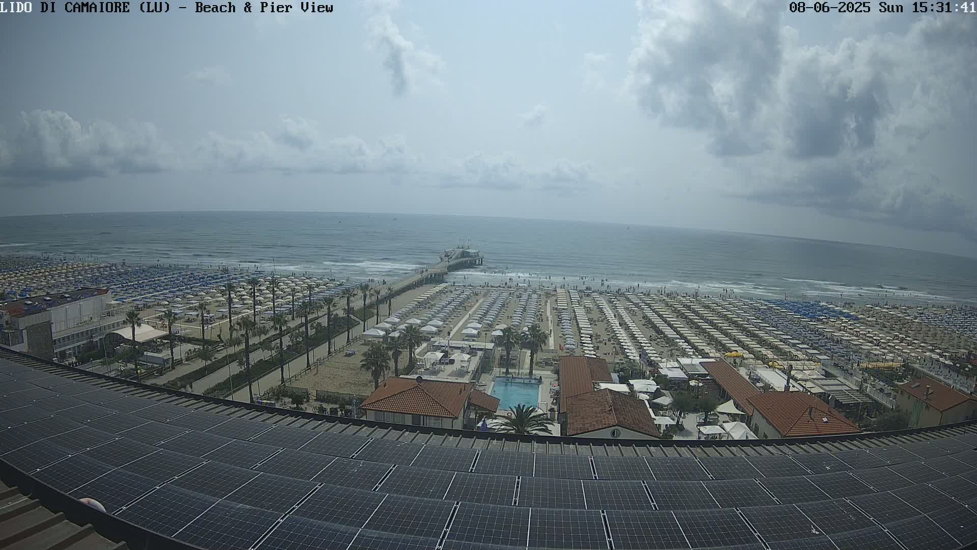 A mostly sunny day overlooks a beach with numerous umbrellas and a pier extending into a wavy ocean, viewed from a rooftop with solar panels.