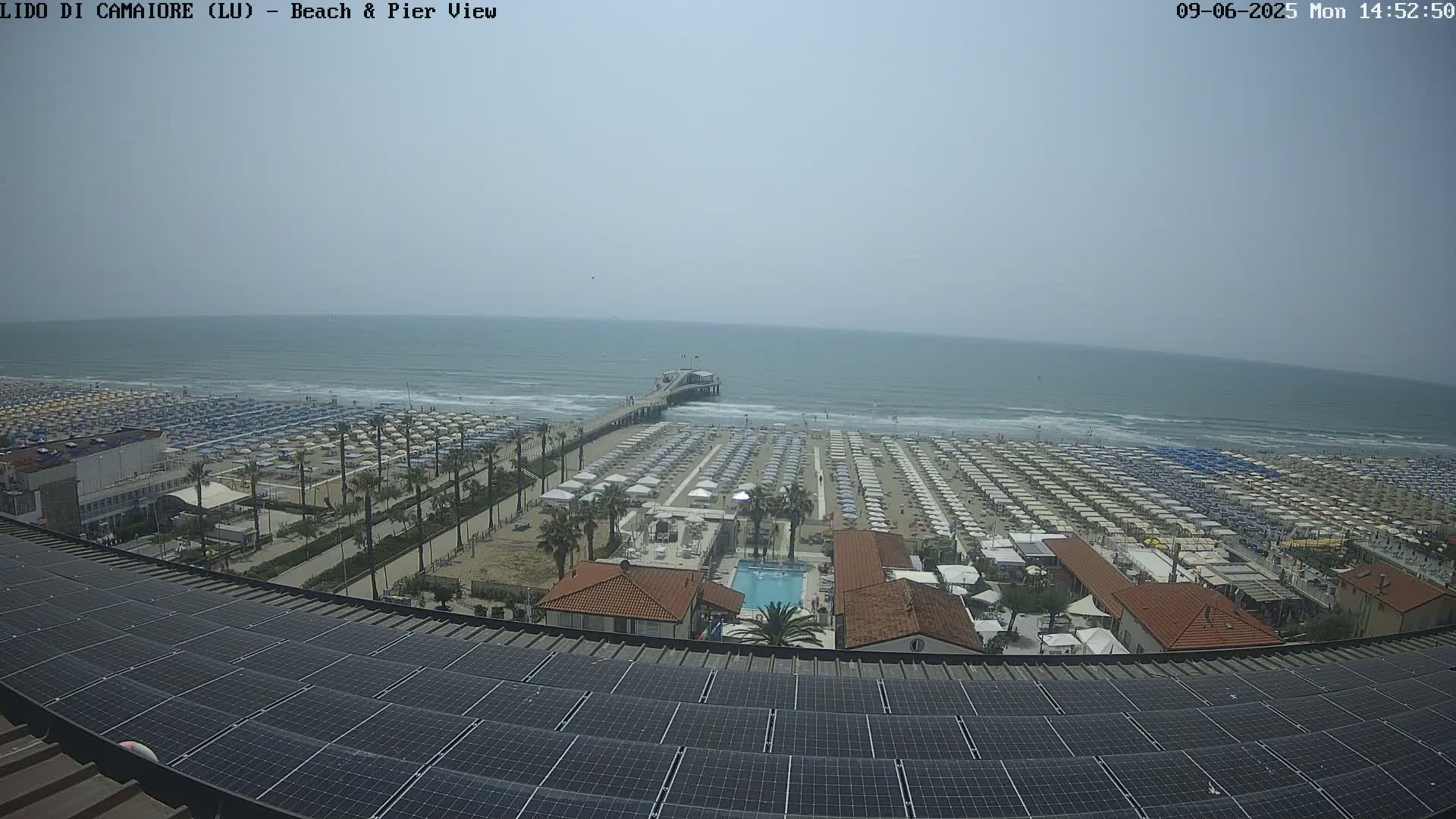 A beach with numerous umbrellas and a pier extends into a calm, hazy ocean, viewed from above a rooftop covered in solar panels.