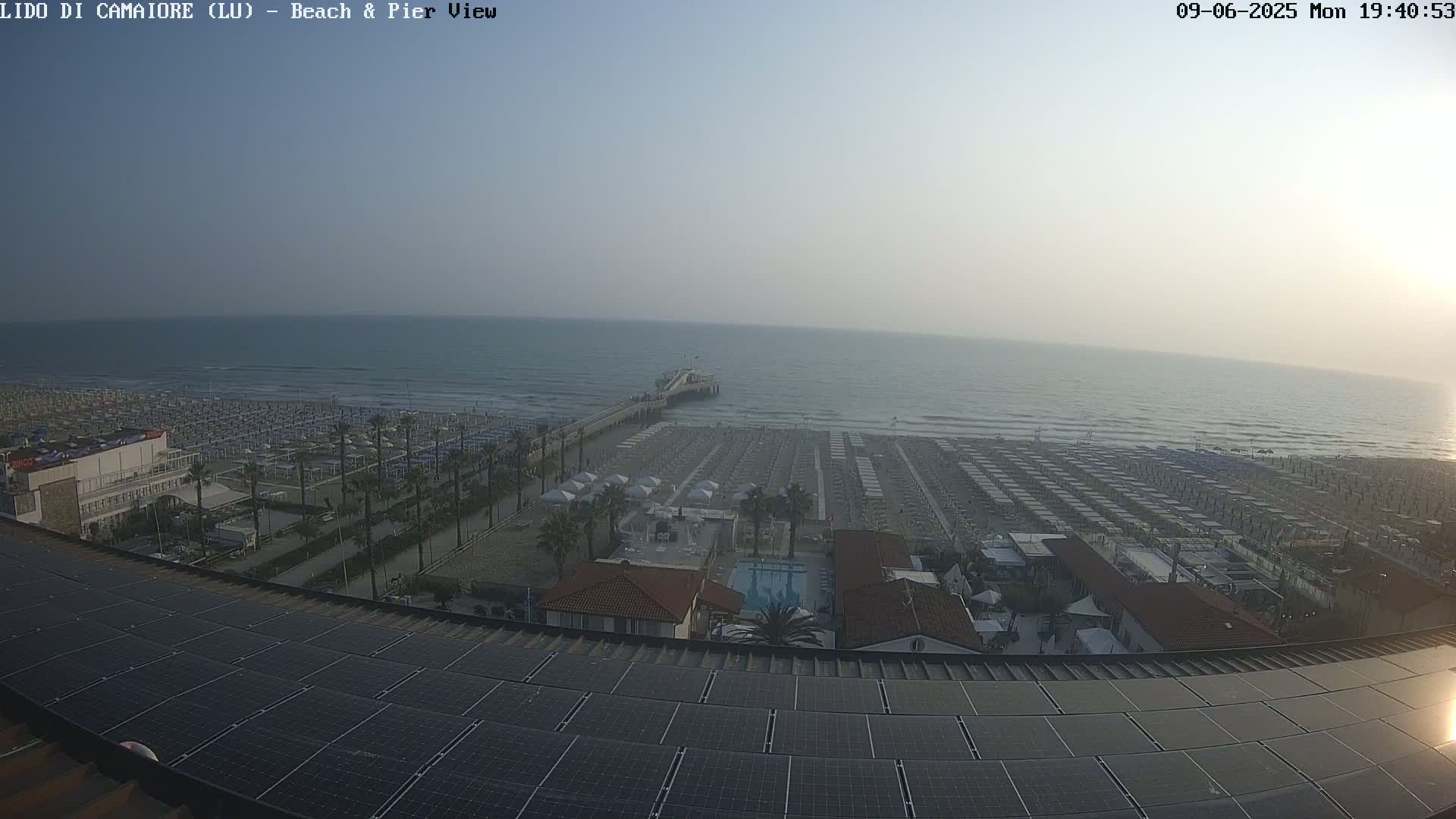 A hazy day shows a beach with numerous umbrellas and lounge chairs, a pier extending into a calm sea, and a solar panel array on a rooftop overlooking the scene.