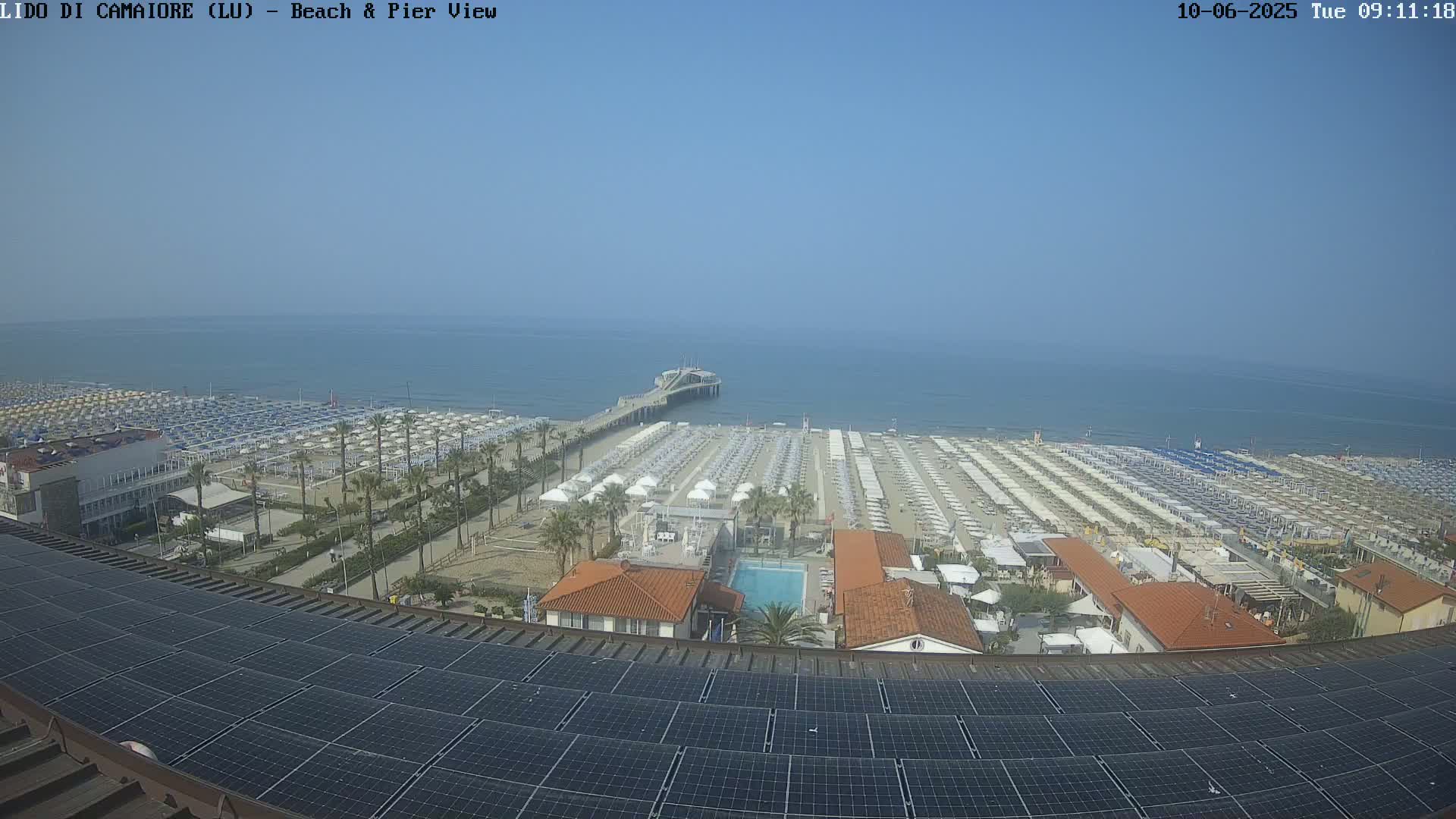 A beach with many umbrellas and a pier extends into a calm, blue sea under a clear sky, viewed from above a rooftop covered in solar panels.