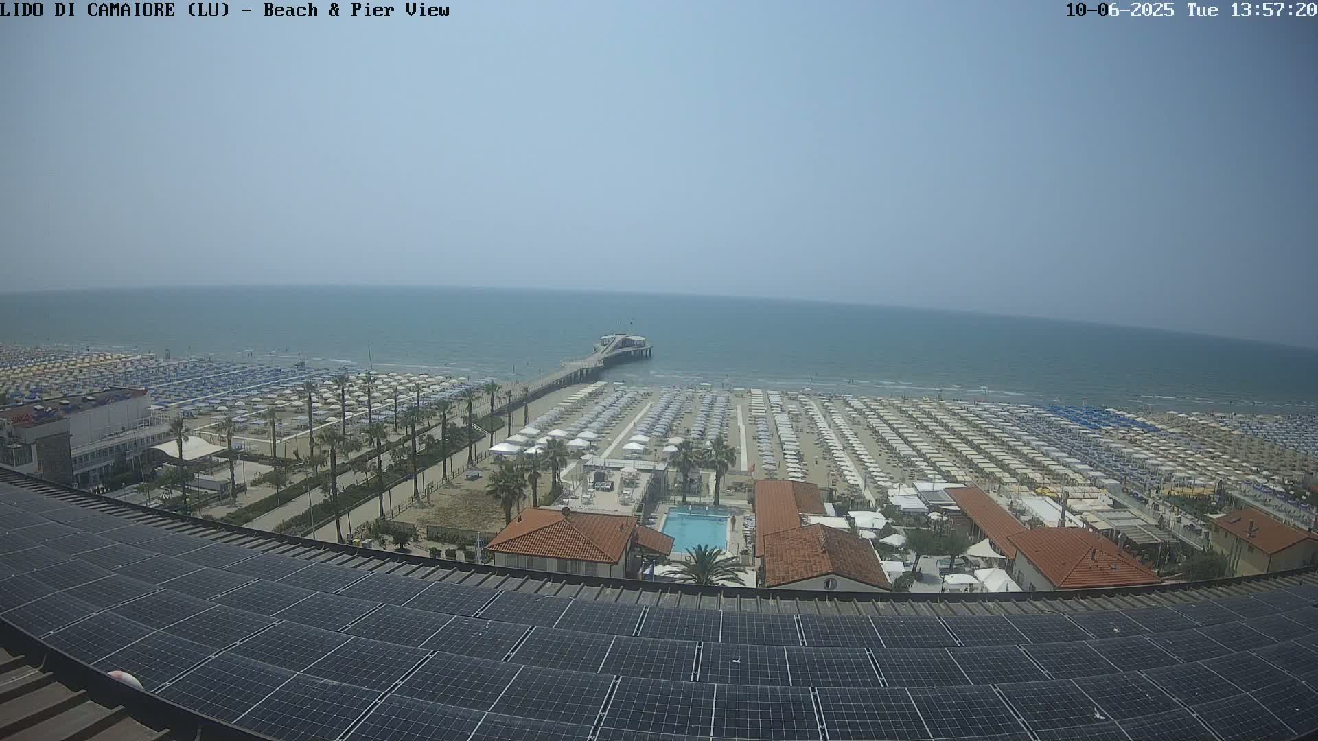 A sunny day at a beach with many umbrellas, a pier, and a building with solar panels on the roof.
