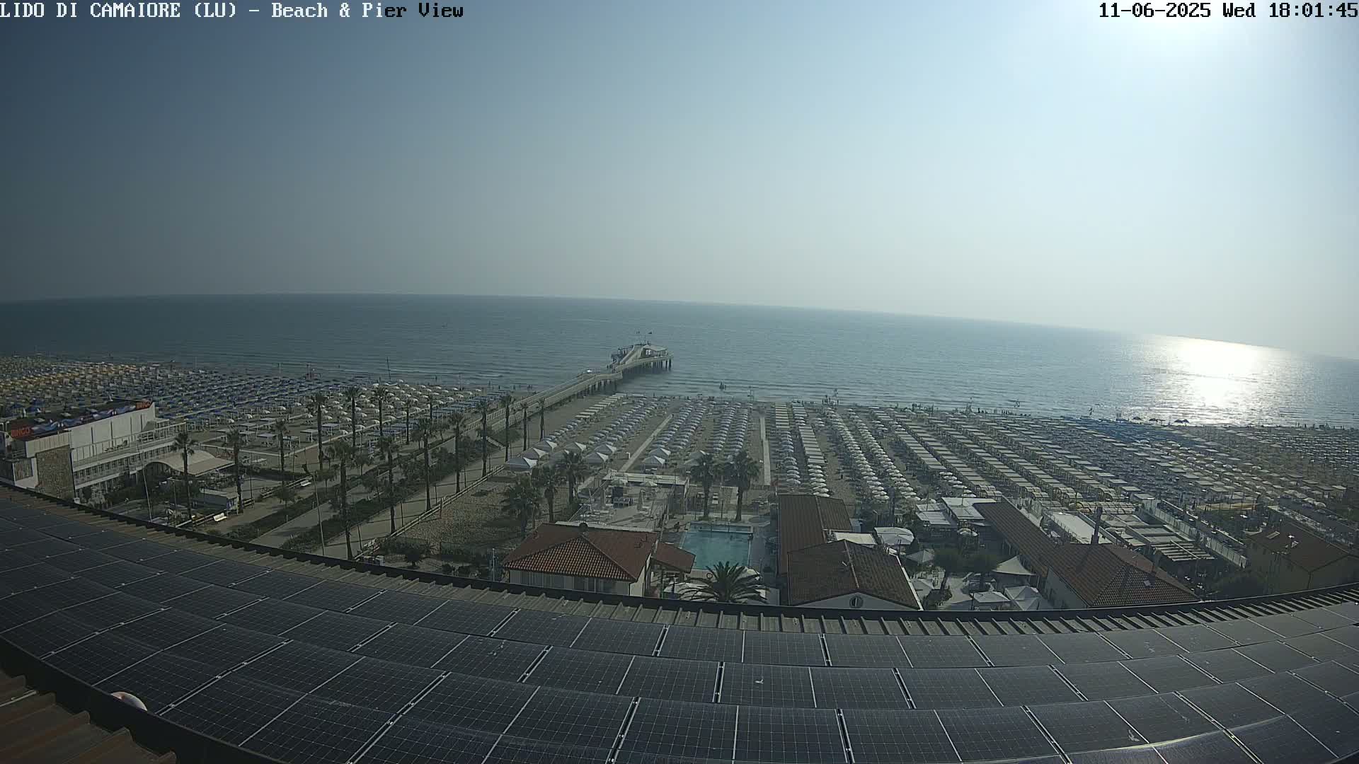 A beach with numerous umbrellas and a pier extends into a calm, sunny ocean, viewed from a rooftop covered in solar panels.