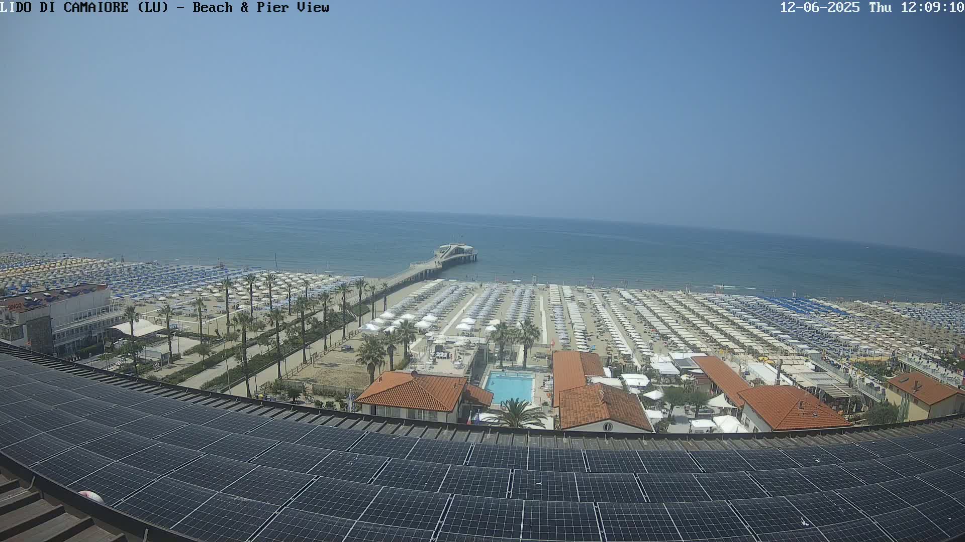 A beach with numerous umbrellas and a pier extending into a calm, blue sea is viewed from above a rooftop covered in solar panels on a sunny day.