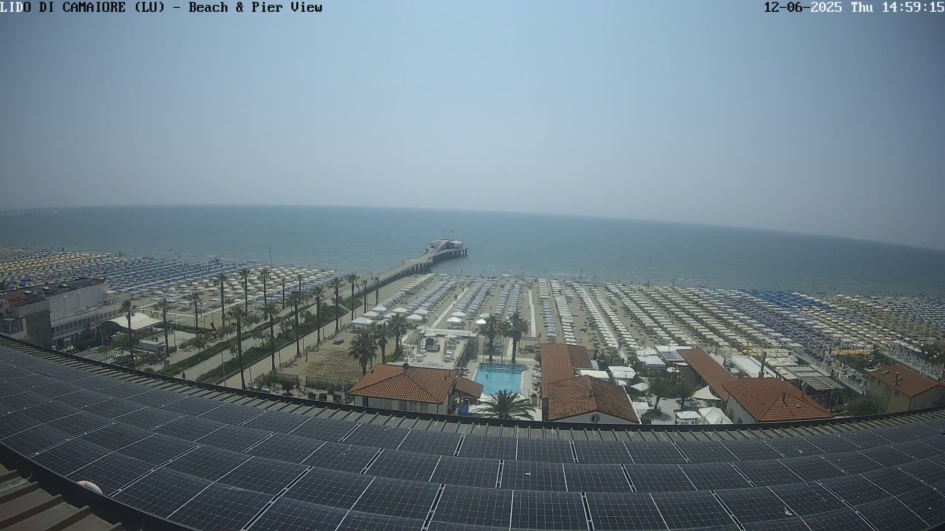 A sunny day overlooking a beach with numerous umbrellas, a pier extending into a calm sea, and a solar panel array in the foreground.