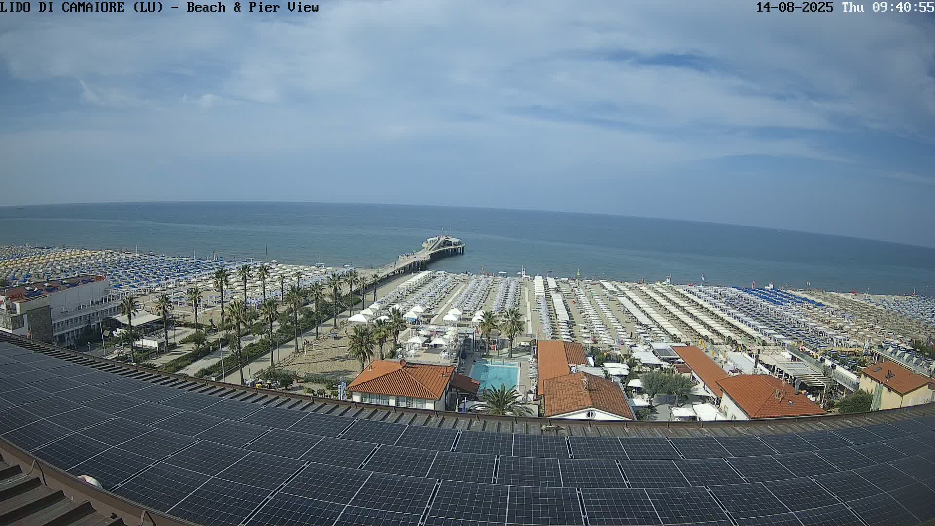 A mostly sunny day overlooking a beach with many umbrellas and a pier extending into a calm sea, viewed from a rooftop covered in solar panels.