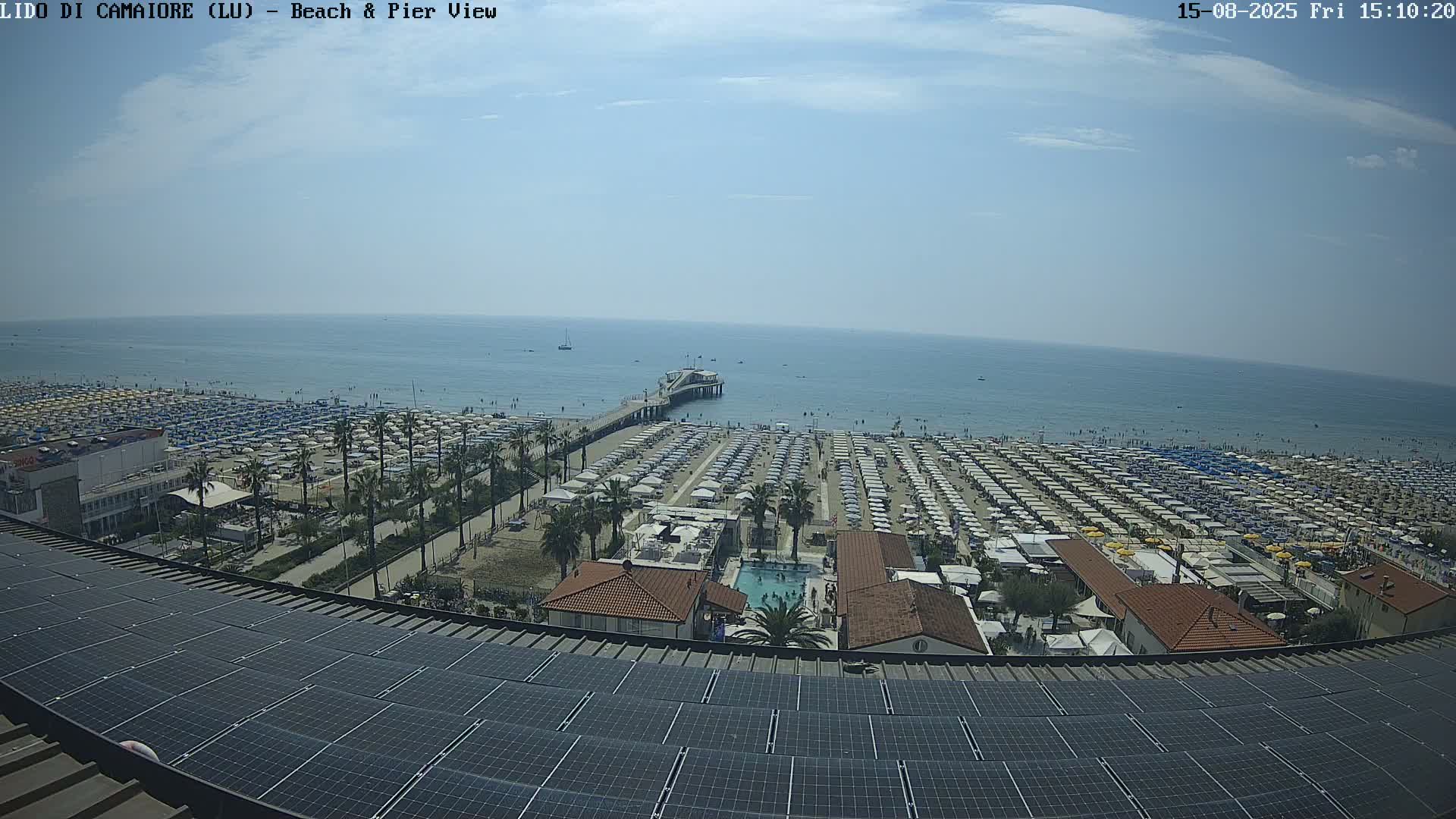 A sunny day shows a beach with many umbrellas and palm trees, a pier extending into a calm sea, and a solar panel array in the foreground.