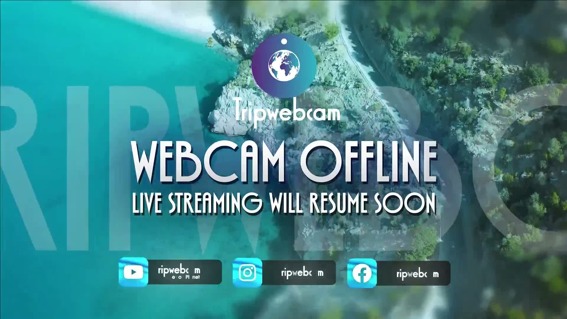An aerial view reveals a tranquil body of turquoise water nestled among lush green, rocky terrain and a winding road, all under clear weather conditions.