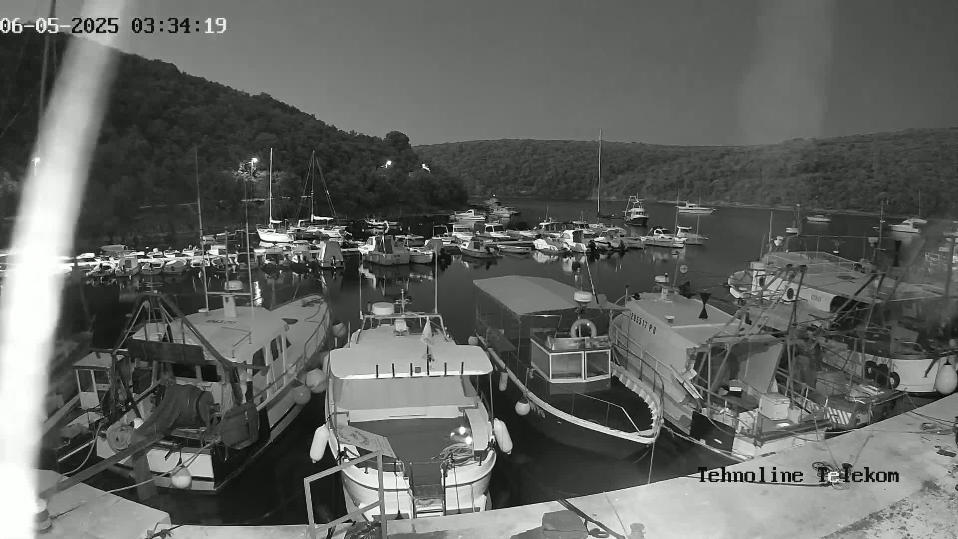 A nighttime, grayscale image shows numerous boats moored in a calm harbor, nestled against a dark, hilly shoreline.