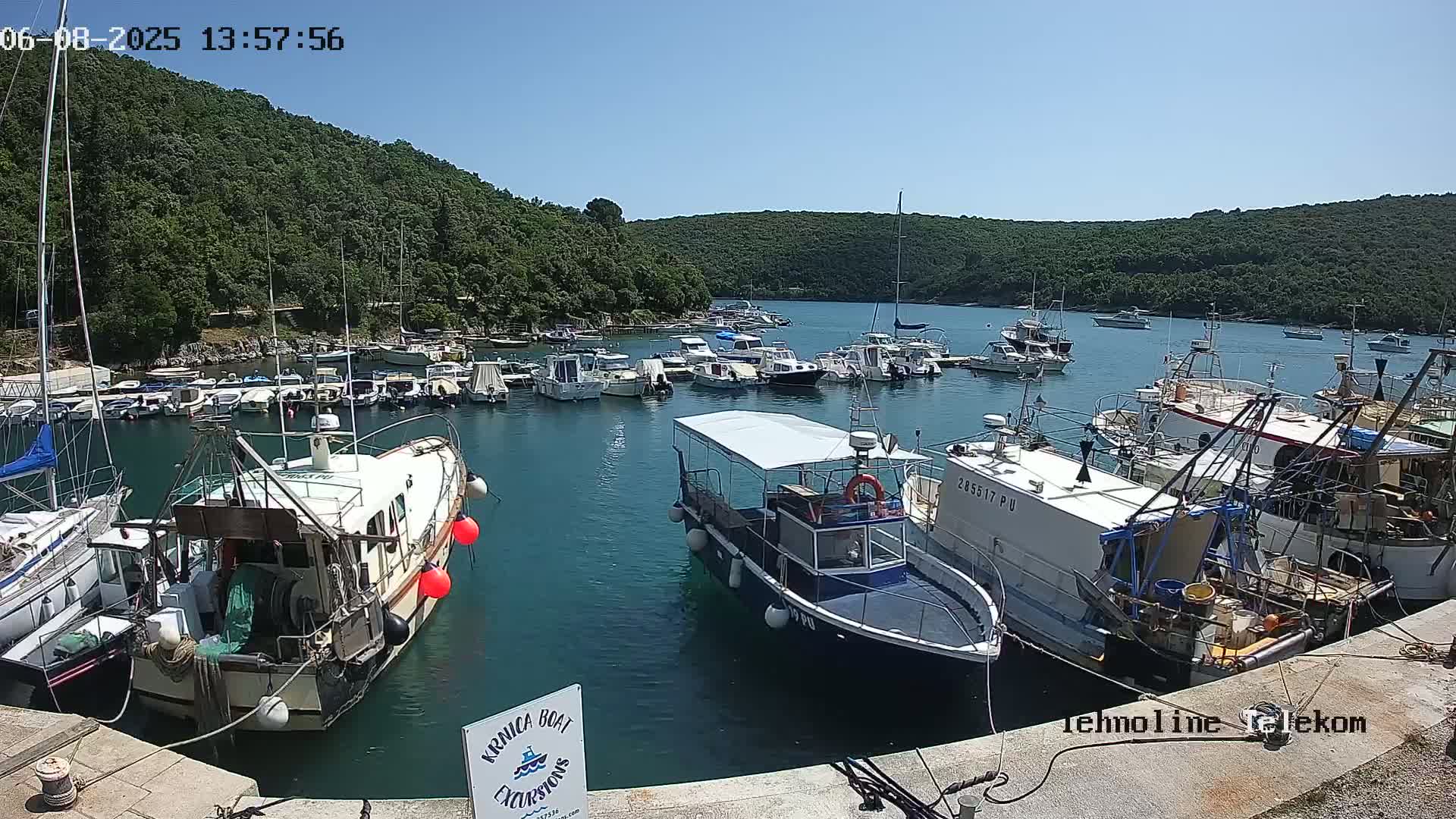 A sunny day shows a calm harbor filled with numerous boats of varying sizes, nestled between green hills.