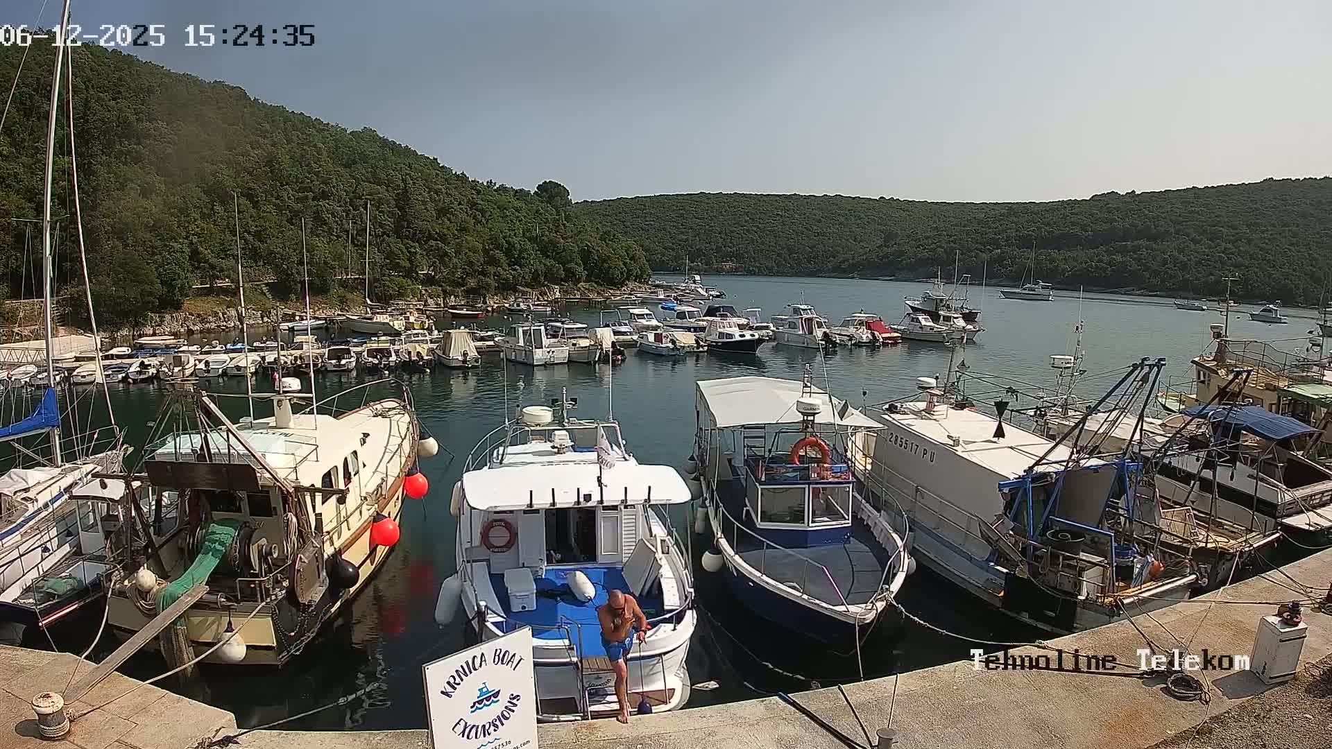 A man stands on the deck of a boat in a harbor filled with numerous other boats, all under a sunny sky, surrounded by lush green hills.