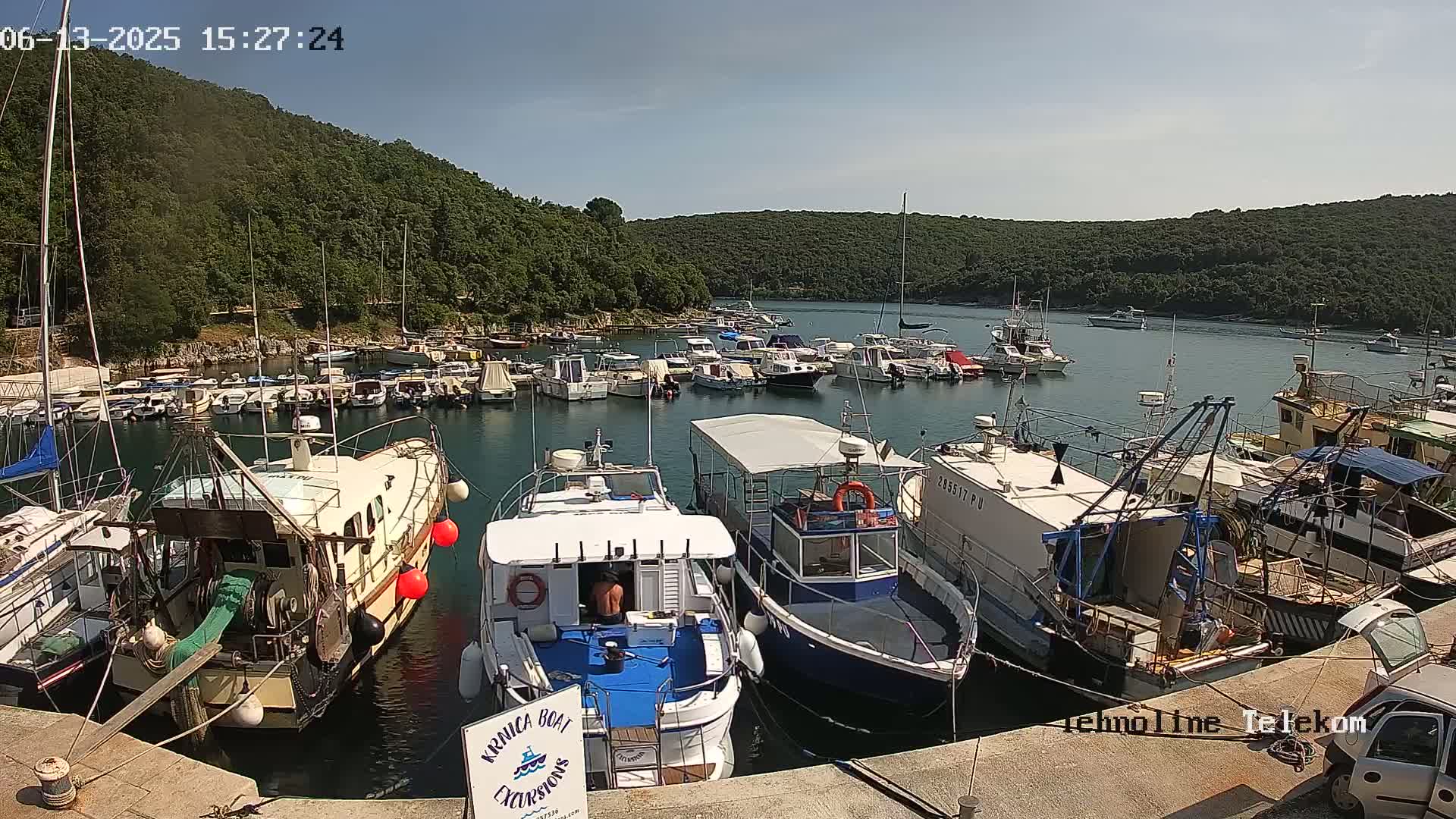 A harbor filled with numerous boats and yachts is nestled in a cove surrounded by lush green hills under a clear, sunny sky.