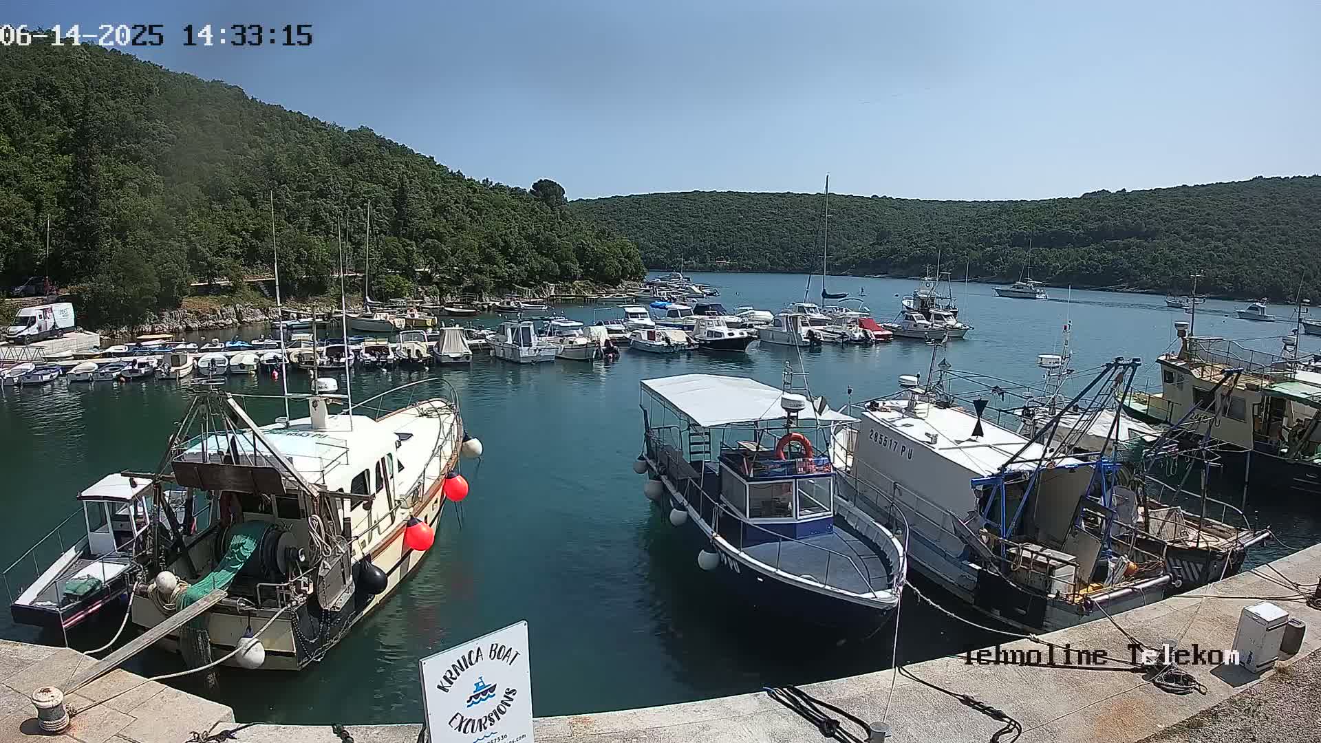 A sunny day shows a calm harbor filled with numerous boats and yachts, surrounded by lush green hills.