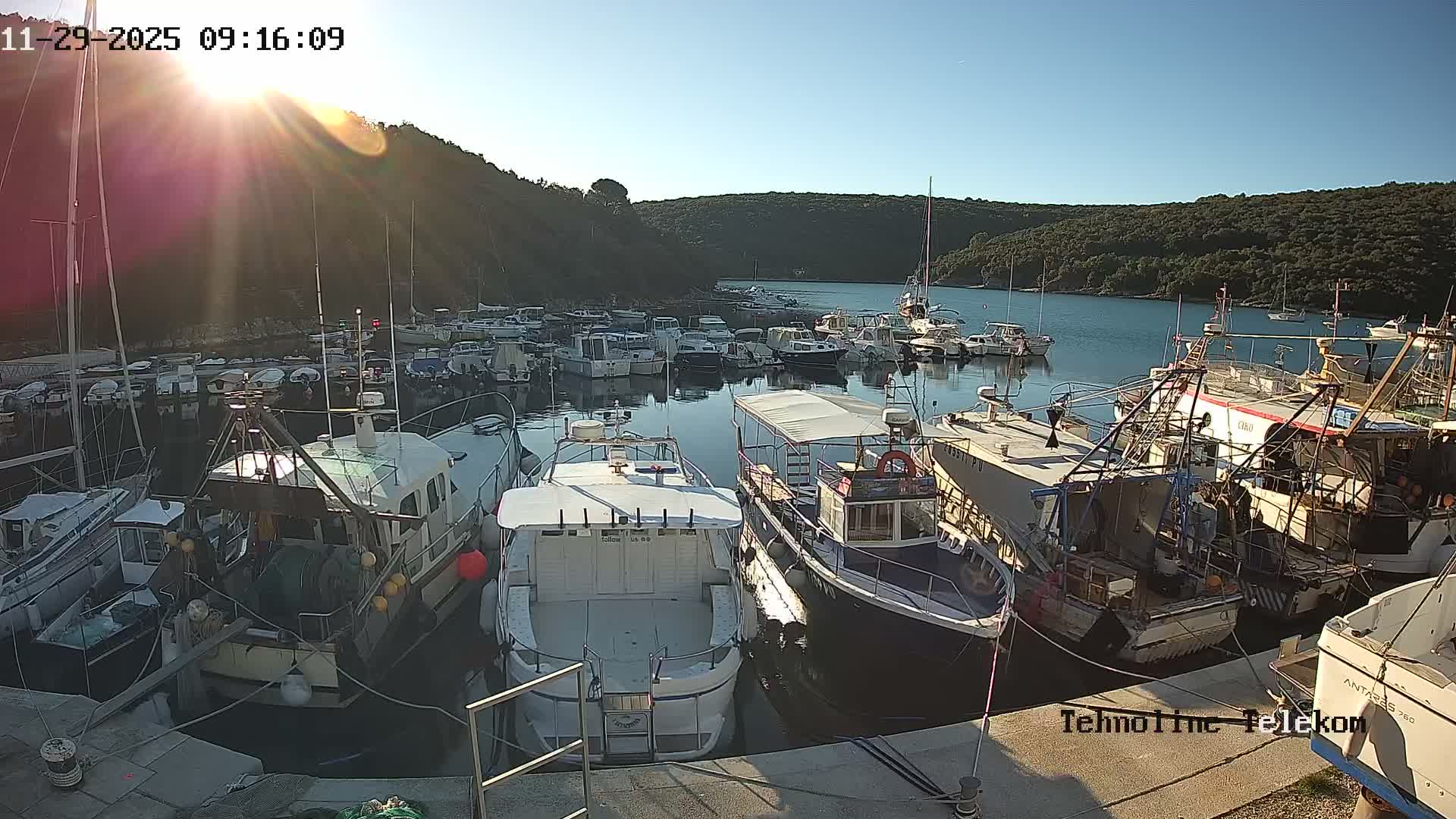 On a bright, sunny morning, numerous boats are moored in a calm, clear bay surrounded by lush, tree-covered hills.