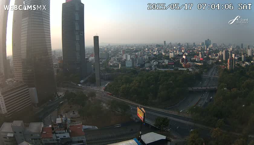 A hazy sunrise illuminates a cityscape of tall buildings and a large highway system surrounded by greenery.