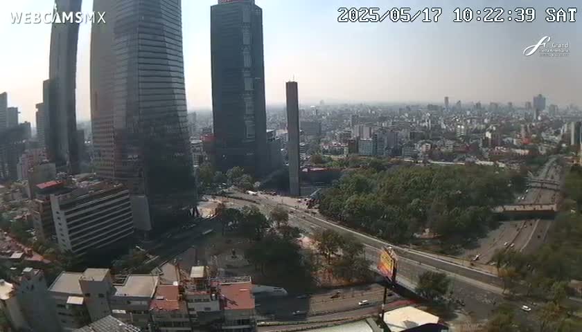 A hazy, high-angle view shows a cityscape with tall buildings and a wide roadway, surrounded by greenery.