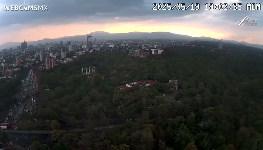 An aerial view at sunset shows a city nestled in a large expanse of green trees and mountains in the background under a cloudy sky.