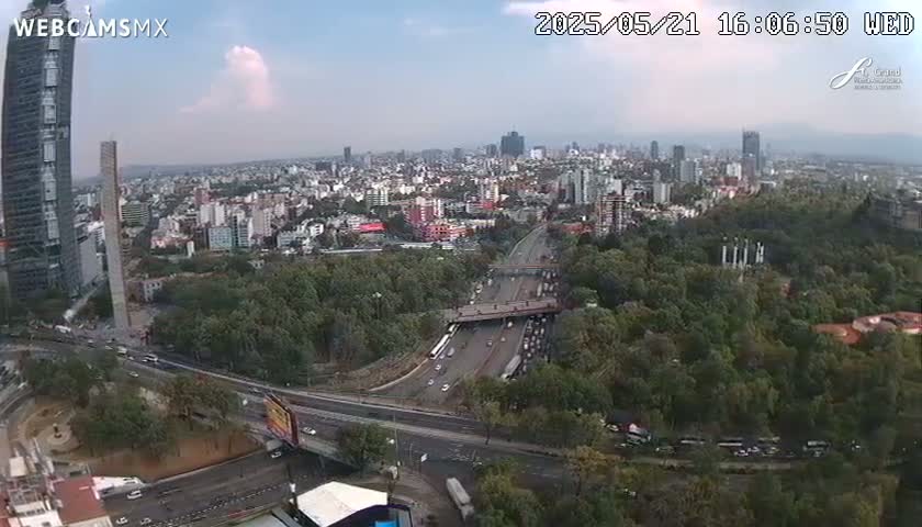 An aerial view of a city on a partly cloudy day, showing a highway weaving through a large park and numerous buildings.