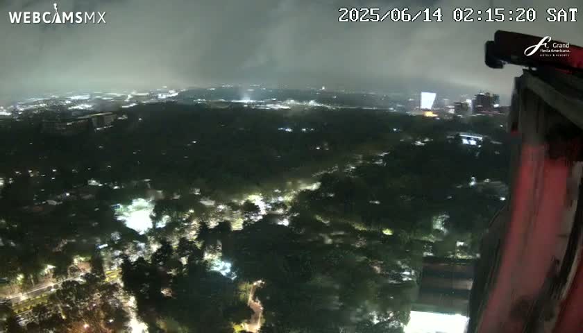 A nighttime, high-angle view shows a sprawling, dark, wooded area speckled with small lights, with a brighter city skyline visible in the distance under an overcast sky.