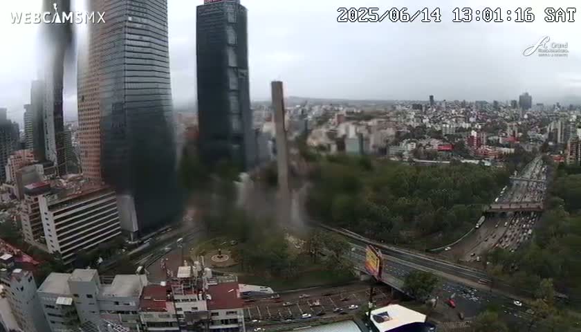 A high-angle, long shot of a city on an overcast day, showing a large highway curving through a green park and past several skyscrapers.