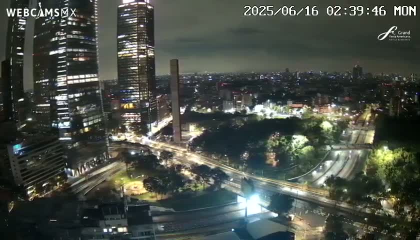 A nighttime aerial view of a city shows several illuminated skyscrapers, roads with moving vehicles, and a park under a cloudy sky.