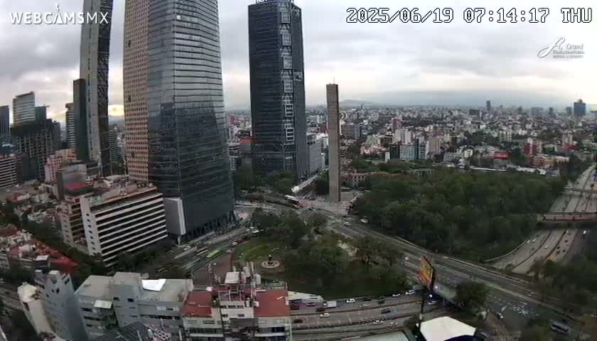 An aerial view of a city with several skyscrapers under a cloudy sky shows roads and green spaces surrounding the buildings.