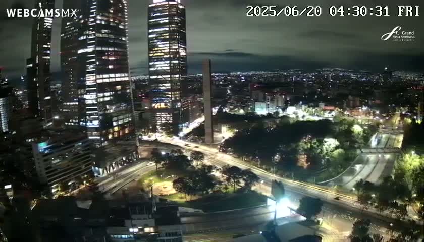 A nighttime aerial view of a city with several skyscrapers, illuminated roads and bridges, and surrounding greenery under a partly cloudy sky.