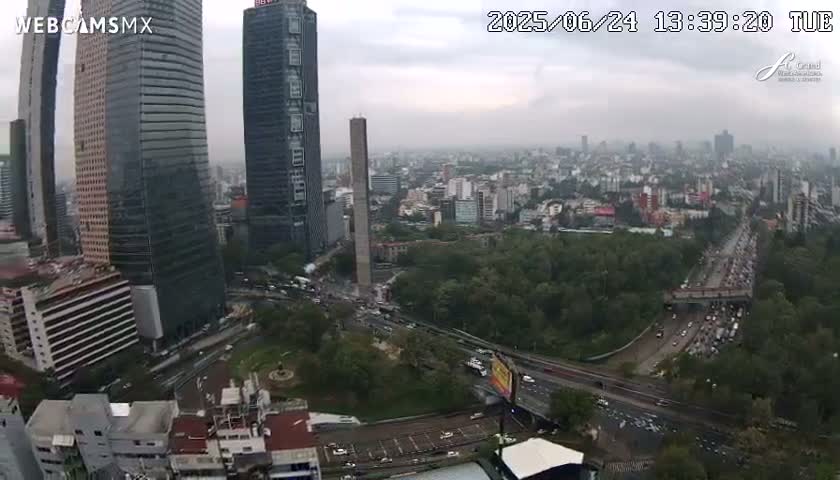 A high-angle, long shot of a city on an overcast day shows tall buildings, a monument, a large expanse of green parkland, and a busy highway.