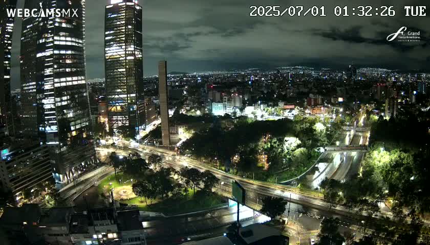 A nighttime aerial view of a city with several skyscrapers, illuminated roads, and green spaces under a cloudy sky.
