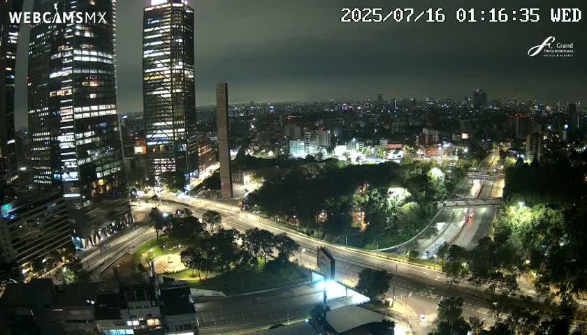 A nighttime aerial view of a city with several tall buildings and a highway system, under a cloudy sky.