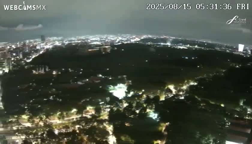 A nighttime aerial view of a city showing illuminated skyscrapers, roadways with moving vehicles, and a park, under a dark, clear sky.